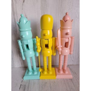 Puede incluir: Tres cascanueces de madera de colores pastel: aguamarina, amarillo y rosa. Cada uno se apoya sobre una base cuadrada y tiene un acabado brillante. El cascanueces amarillo sostiene un bastón, el rosa una espada.