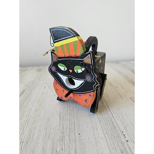 Puede incluir: Una casita de pájaros de madera pintada con un gato negro con un ojo verde y un sombrero naranja y amarillo. La casita tiene un pequeño agujero para que los pájaros puedan entrar. El gato sonríe y lleva una pajarita negra y blanca.