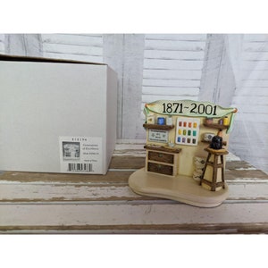 May include: A miniature replica of a shop with a sign that reads "1871-2001". The shop has a wooden facade with a small window and a shelf with colorful paint tubes. The shop is on a cream-colored base.