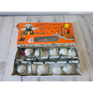 May include: A decorative Halloween 10 light set in its original orange box. The box features a spooky illustration with a haunted house, witch, and jack-o'-lantern. The light set includes white skull and ghost-shaped lights on a green wire.