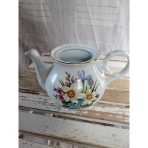 May include: White porcelain teapot with a floral design featuring daffodils, irises, and other flowers in shades of pink, blue, yellow, and purple. The teapot has a gold trim around the rim.