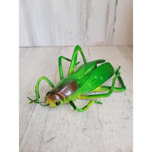 May include: A decorative grasshopper figurine in shades of green, brown, and yellow. The insect has large, detailed wings and long legs, with a detailed head and body. The figurine is made of a durable material and is designed to resemble a real grasshopper.