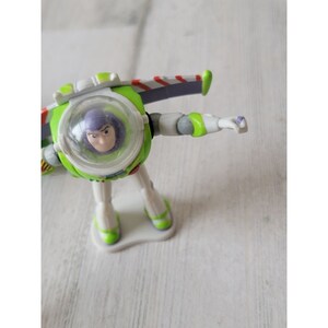 May include: A Buzz Lightyear action figure in a dynamic pose. The toy features a green and white spacesuit with purple accents, a clear helmet, and a determined expression. The figure is on a white base.