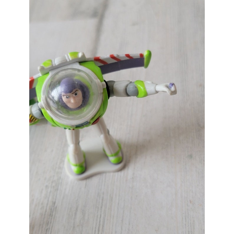 May include: A Buzz Lightyear action figure in a dynamic pose. The toy features a green and white spacesuit with purple accents, a clear helmet, and a determined expression. The figure is on a white base.