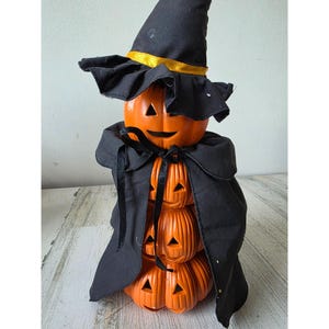 Könnte beinhalten: Eine Halloween-Dekoration mit einem Stapel orangefarbener Kürbisse mit geschnitzten Gesichtern, bedeckt mit einem schwarzen Hexenhut mit goldenem Band und einem schwarzen Umhang. Die Kürbisse sind gestapelt und bilden eine festliche Dekoration.
