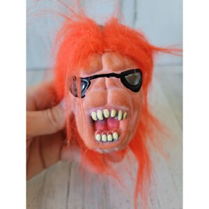 May include: A red haired, orange and pink rubber monster mask with a black eye patch and a toothy grin.