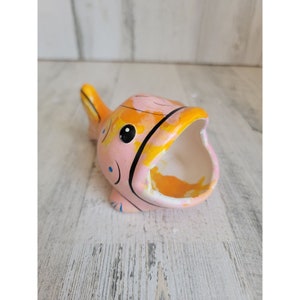 May include: A pink and yellow ceramic fish planter with a black outline and blue details. The fish has an open mouth, creating a planter space.