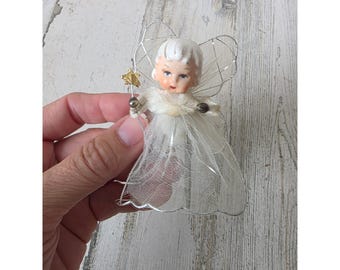 May include: A small angel figurine featuring a white moulded face with painted features, white hair, and a sheer white mesh dress. The angel has silver wire wings, a white faux fur collar, and holds a small gold star wand in one hand.