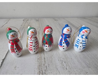 May include: Five snowman ornaments with painted faces, hats, and scarves in red, green, and blue. Each snowman has a gold string for hanging and black dot buttons. The snowmen are on a white wooden surface.