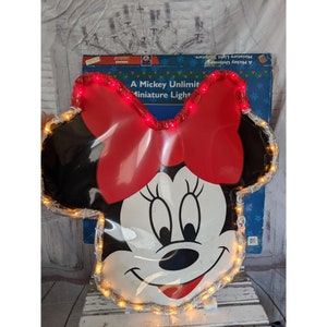 Op de afbeelding: Een grote, rood-zwarte Minnie Mouse lichtdecoratie met witte lampjes die de vorm omlijsten. De strik is rood en het gezicht is wit met zwarte ogen en een zwarte neus. De tekst op de doos luidt "A Mickey Unlimited Miniature Light Sculpture."