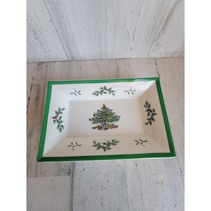 May include: Rectangular white ceramic serving dish with a green border. The dish features a Christmas tree design in the center, surrounded by holly and berry sprigs. A festive holiday serving piece.
