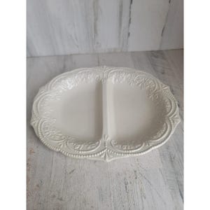 May include: A white, oval-shaped serving platter with two compartments. The platter features an ornate, embossed floral and scroll design around the edges and a central divider. The surface has a glossy finish, and the overall aesthetic is vintage.