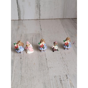 Pode incluir: Uma coleção de figuras em miniatura, incluindo uma princesa e várias figuras em vestidos azuis, segurando petardos. As figuras estão sobre um fundo claro com grão de madeira.