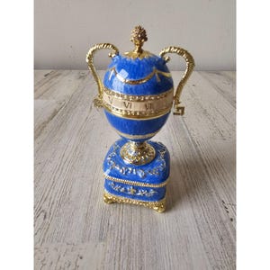 May include: A blue and gold decorative egg-shaped trophy with Roman numerals. The trophy has gold handles and a gold base. The egg-shaped trophy is on a blue square base with gold accents. The top of the trophy is gold with a small gold ornament.