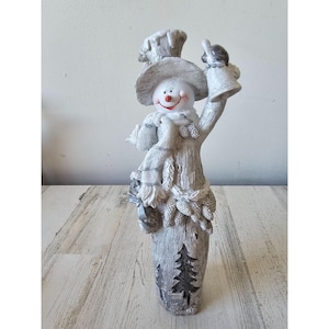 May include: A whimsical snowman figurine with a hat, scarf, and a bell. The snowman has a smiling face with a red nose and is holding a bell in one hand. The base of the figurine has a tree design. The color scheme is primarily white and gray.