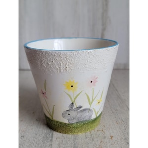 May include: A white ceramic flower pot with a textured surface and a light blue rim. The pot features a hand-painted design of a gray bunny, yellow and pink flowers, and green grass.