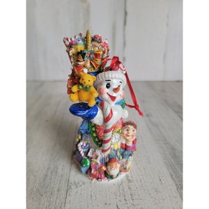 Op de afbeelding: Een witte sneeuwpop kerstversiering met een rood-witte snoepstok, een gele teddybeer en een notenkraker in een mandje bovenop. De versiering is versierd met glitter en kleurrijke details.