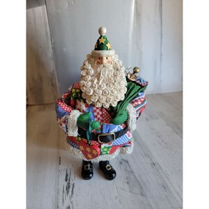 Peut inclure: Une figurine de Père Noël en patchwork avec une barbe blanche et un chapeau vert avec des étoiles dorées. Il tient un sac vert avec une cloche dorée et un cœur rouge avec des étoiles dorées.