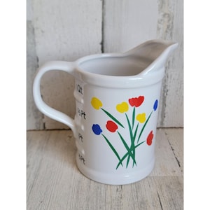 Könnte beinhalten: Weißer Keramik-Krug mit Blumenmuster. Der Krug hat einen Henkel und Markierungen zum Messen in Quarts und Pints.