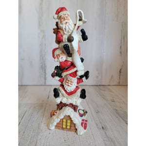 May include: A stack of three Father Christmas figurines climbing a chimney. The Father Christmas figurines are made of resin and are painted in red, white, and black. The chimney is made of white resin and has a snowy roof.