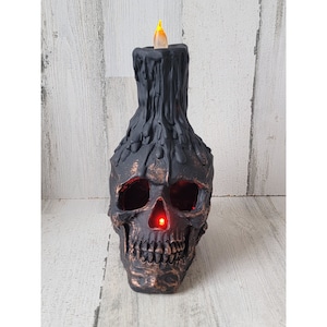 May include: Black skull candle holder with a dripping wax effect. The candle is lit and has a warm orange glow.