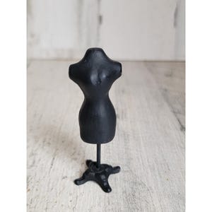 May include: A small, black dress form with a detailed bodice and a flared skirt. The miniature mannequin is mounted on a slender metal rod, which is attached to a decorative, star-shaped base. The dress form is a classic sewing and crafting accessory.