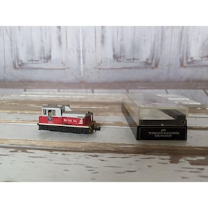 May include: A red and silver toy train engine with the text "Burlington  704" on the side. The engine is in a clear plastic display case with a black base. The base has the text "4791 "Plymouth 0-6-0 Diesel Burlington" on it.