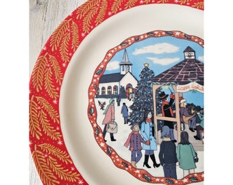 May include: A white ceramic plate with a red and gold border featuring a winter scene with a church, a Christmas tree, and people. The text "Happy Holidays" is visible on the plate.