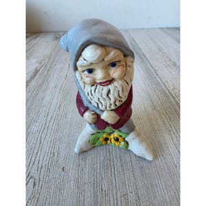 May include: A ceramic garden gnome figurine with a gray hat, red jacket, and white beard. The gnome is holding a gray tool and is perched on a base with yellow flowers and green leaves.