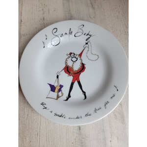 May include: White ceramic plate with a hand-painted illustration of Santa Claus carrying a bag of gifts. The plate has the text "Santa Baby Slip a sable under the tree for me".