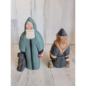 May include: Two wooden figurines of people dressed in winter clothing. The figurine on the left is wearing a blue hooded coat and holding a small dog. The figurine on the right is wearing a black hooded coat and has a white beard.