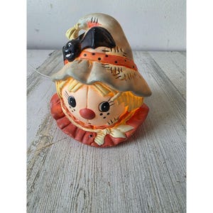 Könnte beinhalten: Eine keramische Halloween-Lampe in Form einer Vogelscheuche. Die Lampe zeigt einen Vogelscheuchenkopf mit Strohhut, roter Nase und rotem Kragen. Die Lampe leuchtet von innen und erzeugt einen warmen Schein. Auf dem Hut der Vogelscheuche sitzt eine schwarze Krähe.