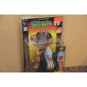 May include: A Spawn action figure of the character Clown, in a black and white striped shirt, black jacket, and white mask. The figure is holding a mallet in its right hand. The figure is packaged in a plastic blister pack with a comic book. The comic book cover features the Spawn logo and the character Clown.