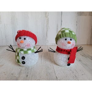 May include: Two decorative snowman figures. One snowman wears a red knit hat and a green and white plaid scarf. The other snowman wears a green and white plaid knit hat and a red scarf. Both have black twig arms and button eyes.