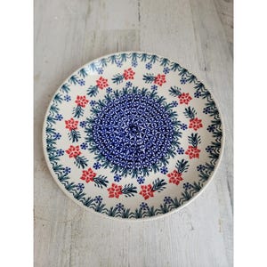 May include: A round, white ceramic plate with a detailed floral design. The plate features a central blue pattern surrounded by red flowers and green leaves. The rim of the plate has a repeating pattern of green leaves.