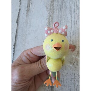 May include: A yellow chick ornament with a glittery finish, featuring a pink bow with white polka dots on its head. The chick has an orange beak, small blue eyes, and orange feet. A small pink loop is attached to the top for hanging purposes.