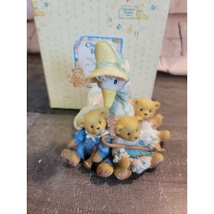 May include: A white goose figurine wearing a yellow and white hat and a blue dress, holding a walking stick. Three teddy bear figurines are sitting on the goose's back and legs. The teddy bears are wearing blue and white clothing.