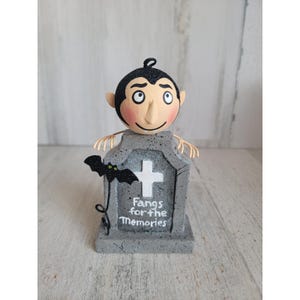 Könnte beinhalten: Eine Halloween-Figur mit einem Cartoon-Vampir auf einem grauen Grabstein. Der Grabstein hat ein weißes Kreuz und den Text "Fangs for the Memories". Eine schwarze Fledermaus mit grünen Glitzerakzenten ist am Grabstein befestigt.