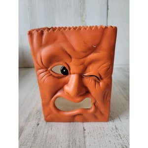 May include: Orange ceramic Halloween candle holder shaped like a face with one eye open and one eye closed. The face is frowning and has a large mouth.