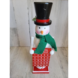 Può includere: Un calendario natalizio a forma di pupazzo di neve. Il pupazzo di neve ha un cappello a cilindro nero, una sciarpa verde e un calendario rosso con i numeri da 1 a 25. Il viso del pupazzo di neve è dipinto con guance rosee e un naso a carota.
