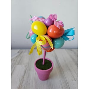 May include: A pink ceramic pot with green grass inside holds a decorative Easter tree. The tree is made of a stick with colorful Easter eggs and pink and blue ribbons attached.