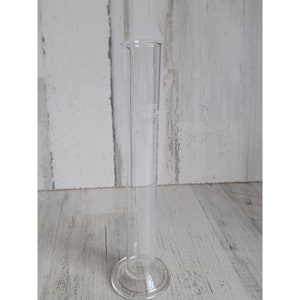 May include: A clear glass graduated cylinder with a capacity of 100 milliliters. The cylinder has markings for measuring volumes in milliliters.
