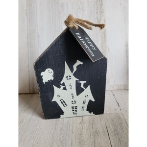 May include: Black wooden Halloween decoration shaped like a house, featuring a white haunted house silhouette and a ghost. A tag with "Happy Halloween" hangs from the top with twine. The house is set against a light wood background.