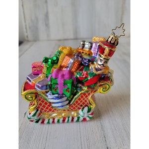 May include: A glass Christmas ornament depicting a nutcracker in a red and gold sleigh filled with colorful presents. The ornament has a gold star on top.