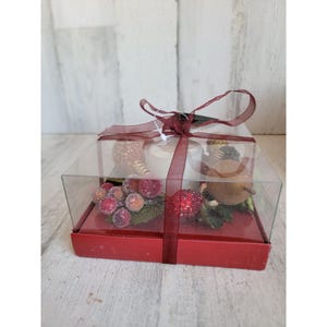 May include: A clear plastic box with a red lid and a red ribbon tied around it. Inside the box are two white candles, a gold and brown decorative piece, and a red and green berry sprig.