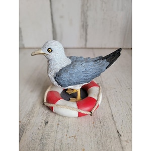 Puede incluir: Una figurita de gaviota blanca y gris posada sobre un salvavidas rojo y blanco. La figurita está hecha de resina y tiene un aspecto realista.