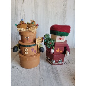 Puede incluir: Dos decoraciones navideñas hechas a mano. Una es una pila de cuatro cajas con una cara de reno marrón y un patrón de cuadros verde y blanco. La otra es una figura de Papá Noel roja y verde que sostiene un pequeño árbol de Navidad. El letrero dice "Feliz Navidad".