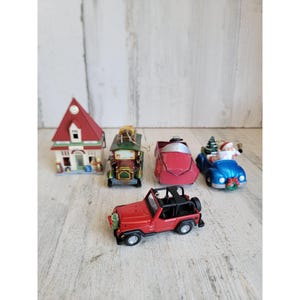 May include: A collection of Christmas-themed miniature vehicles and a house ornament. The ornaments include a red Jeep, a blue car with Santa, a red car, a green and gold antique car, and a house with a red roof.