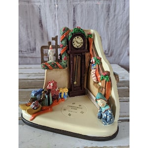 May include: A ceramic Christmas scene figurine with a clock, presents, and a small elf. The figurine is painted in bright colors and has a whimsical feel. The base of the figurine reads "Christmas Time Hum 2106".