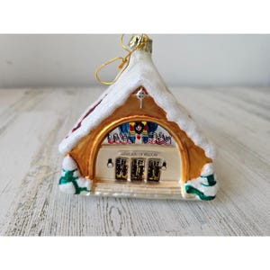 Puede incluir: Un adorno de Navidad de vidrio con forma de iglesia con un techo blanco nevado, adornos dorados y una ventana de vidriera con el texto "MART'S SEAT OF WISDOM".